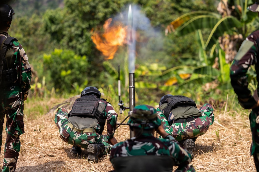 Military personnel in camouflage uniforms and tactical vests are operating a mortar system in an outdoor field environment. A projectile has just been fired, with a visible flame and smoke trail above the mortar tube. The scene appears to be a military training exercise or live-fire demonstration.