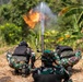 Military personnel in camouflage uniforms and tactical vests are operating a mortar system in an outdoor field environment. A projectile has just been fired, with a visible flame and smoke trail above the mortar tube. The scene appears to be a military training exercise or live-fire demonstration.