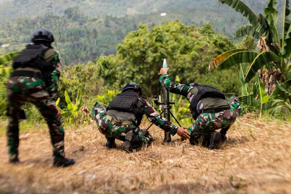 Three soldiers in camouflage uniforms and tactical vests are operating a mortar in an outdoor, vegetated hillside environment. Two soldiers are crouched around the mortar tube while one holds what appears to be a mortar round above the tube. A third soldier stands nearby in a blurred foreground position.