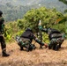 Three soldiers in camouflage uniforms and tactical vests are operating a mortar in an outdoor, vegetated hillside environment. Two soldiers are crouched around the mortar tube while one holds what appears to be a mortar round above the tube. A third soldier stands nearby in a blurred foreground position.