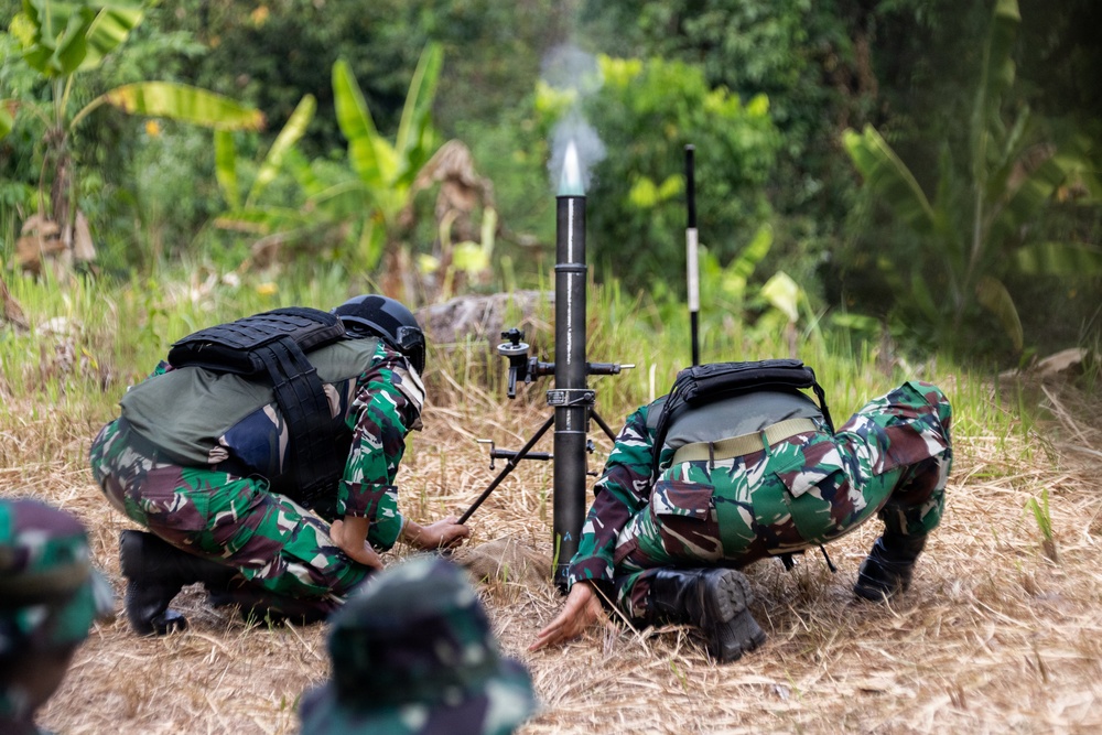 Two soldiers in camouflage uniforms are operating a mortar system in a field environment surrounded by tropical vegetation. Smoke is visible at the top of the mortar tube, indicating a recent or ongoing firing. The soldiers are wearing tactical gear including helmets and body armor/backpacks.