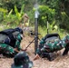 Two soldiers in camouflage uniforms are operating a mortar system in a field environment surrounded by tropical vegetation. Smoke is visible at the top of the mortar tube, indicating a recent or ongoing firing. The soldiers are wearing tactical gear including helmets and body armor/backpacks.