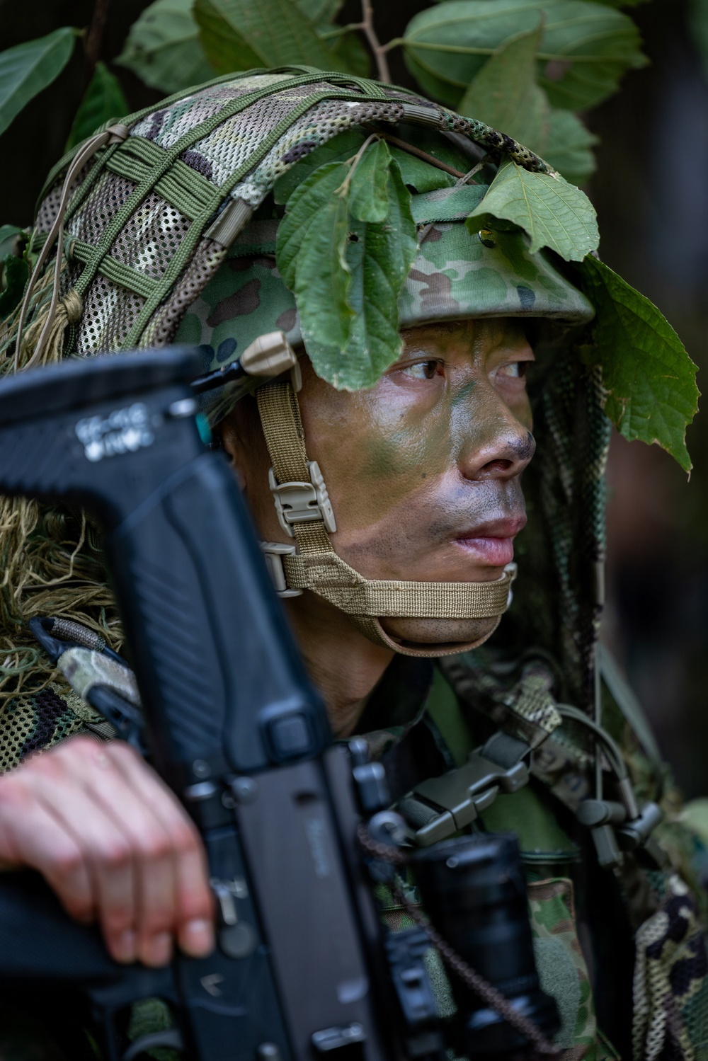 A soldier in full camouflage gear is positioned in a forested environment, holding a rifle and scanning surroundings. The individual has green and black face paint applied and wears a tactical helmet adorned with natural foliage for concealment. The image appears to depict a military field exercise or patrol operation in a jungle or dense woodland setting.