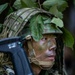 A soldier in full camouflage gear is positioned in a forested environment, holding a rifle and scanning surroundings. The individual has green and black face paint applied and wears a tactical helmet adorned with natural foliage for concealment. The image appears to depict a military field exercise or patrol operation in a jungle or dense woodland setting.