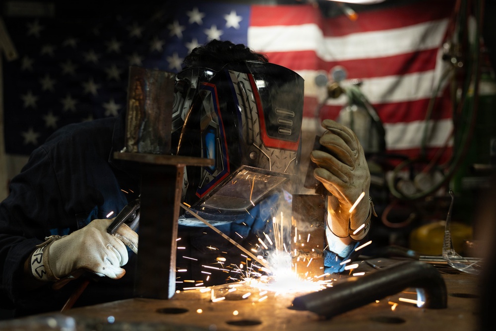 A welder is actively welding metal components in what appears to be a workshop or garage setting. The worker is wearing full protective gear including a decorative welding helmet and leather gloves, with bright sparks flying from the weld point. An American flag is prominently displayed in the background, suggesting a patriotic or American-made work environment.