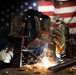 A welder is actively welding metal components in what appears to be a workshop or garage setting. The worker is wearing full protective gear including a decorative welding helmet and leather gloves, with bright sparks flying from the weld point. An American flag is prominently displayed in the background, suggesting a patriotic or American-made work environment.