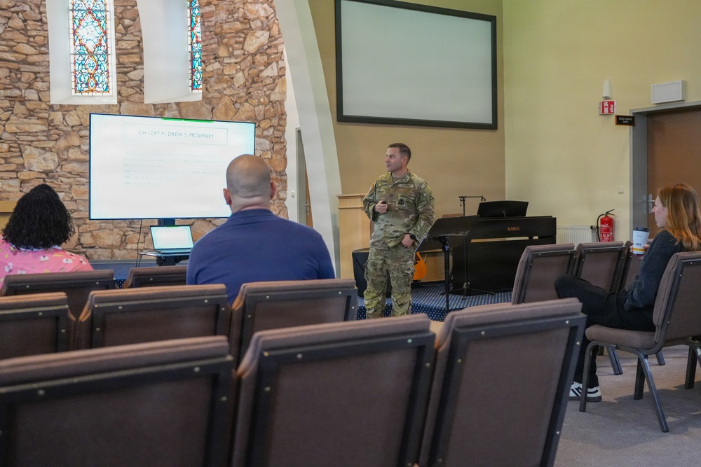 A military personnel in camouflage uniform is presenting to a small audience in what appears to be a chapel or religious facility converted for briefing use. A display screen shows text referencing 'CH (CPT) Drew S. McGinley.' Three civilians are seated in the audience area, with many chairs unoccupied.