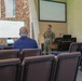 A military personnel in camouflage uniform is presenting to a small audience in what appears to be a chapel or religious facility converted for briefing use. A display screen shows text referencing 'CH (CPT) Drew S. McGinley.' Three civilians are seated in the audience area, with many chairs unoccupied.