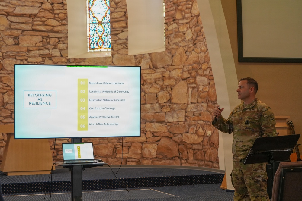 A U.S. Army soldier in OCP (Operational Camouflage Pattern) uniform is delivering a presentation titled 'Belonging as Resilience' in what appears to be a military chapel setting. The presentation slide outlines six topics related to loneliness, community, and resilience. The venue features stone walls and stained glass windows characteristic of a chapel facility.
