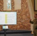 A U.S. Army soldier in OCP (Operational Camouflage Pattern) uniform is delivering a presentation titled 'Belonging as Resilience' in what appears to be a military chapel setting. The presentation slide outlines six topics related to loneliness, community, and resilience. The venue features stone walls and stained glass windows characteristic of a chapel facility.