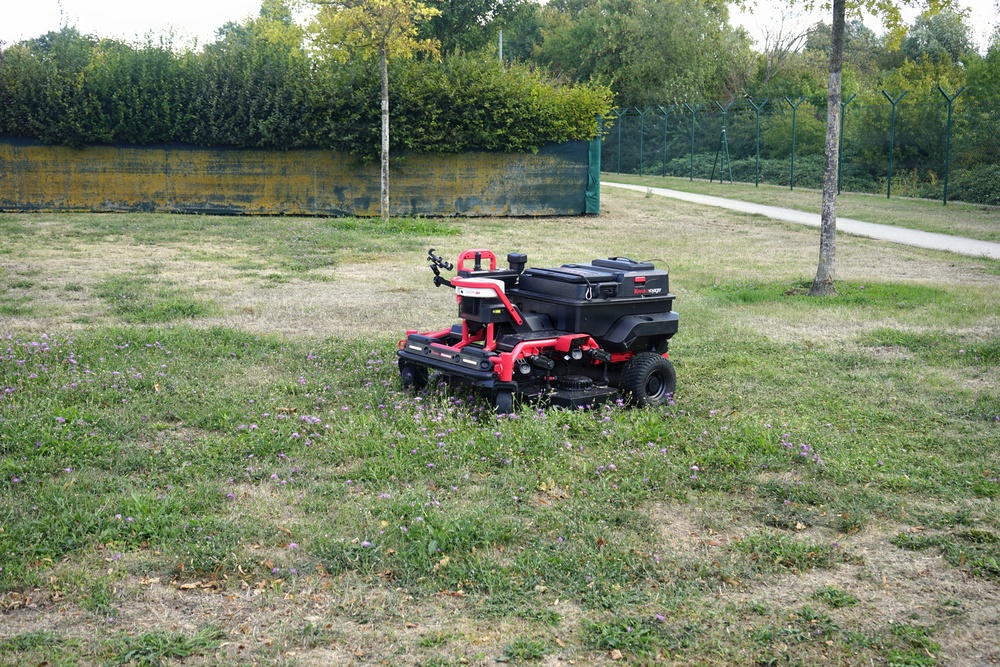 A red and black autonomous or remote-controlled riding lawn mower is positioned on a partially mowed grass area with scattered small purple wildflowers. The setting appears to be a public park or institutional grounds, bordered by a hedge, a yellow-painted wall, and a green metal fence. The grass shows uneven growth with some dry patches, suggesting active maintenance work.
