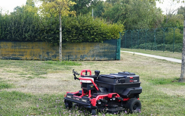 A red and black autonomous or remote-controlled riding lawn mower is positioned on a partially mowed grass area with scattered small purple wildflowers. The setting appears to be a public park or institutional grounds, bordered by a hedge, a yellow-painted wall, and a green metal fence. The grass shows uneven growth with some dry patches, suggesting active maintenance work.