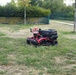 A red and black autonomous or remote-controlled riding lawn mower is positioned on a partially mowed grass area with scattered small purple wildflowers. The setting appears to be a public park or institutional grounds, bordered by a hedge, a yellow-painted wall, and a green metal fence. The grass shows uneven growth with some dry patches, suggesting active maintenance work.