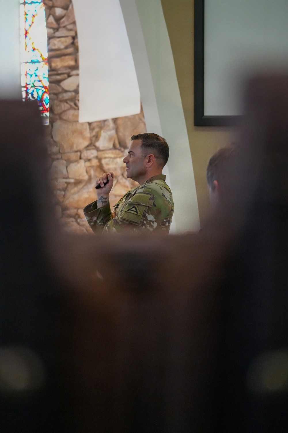 A U.S. Army soldier in Operational Camouflage Pattern (OCP) uniform is speaking or presenting inside what appears to be a military chapel, identifiable by the stained glass window and stone arch architecture. The soldier holds what appears to be a microphone or clicker and is facing an audience. The photo is taken from behind seated audience members, creating a shallow depth-of-field effect with blurred foreground figures.