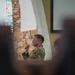 A U.S. Army soldier in Operational Camouflage Pattern (OCP) uniform is speaking or presenting inside what appears to be a military chapel, identifiable by the stained glass window and stone arch architecture. The soldier holds what appears to be a microphone or clicker and is facing an audience. The photo is taken from behind seated audience members, creating a shallow depth-of-field effect with blurred foreground figures.