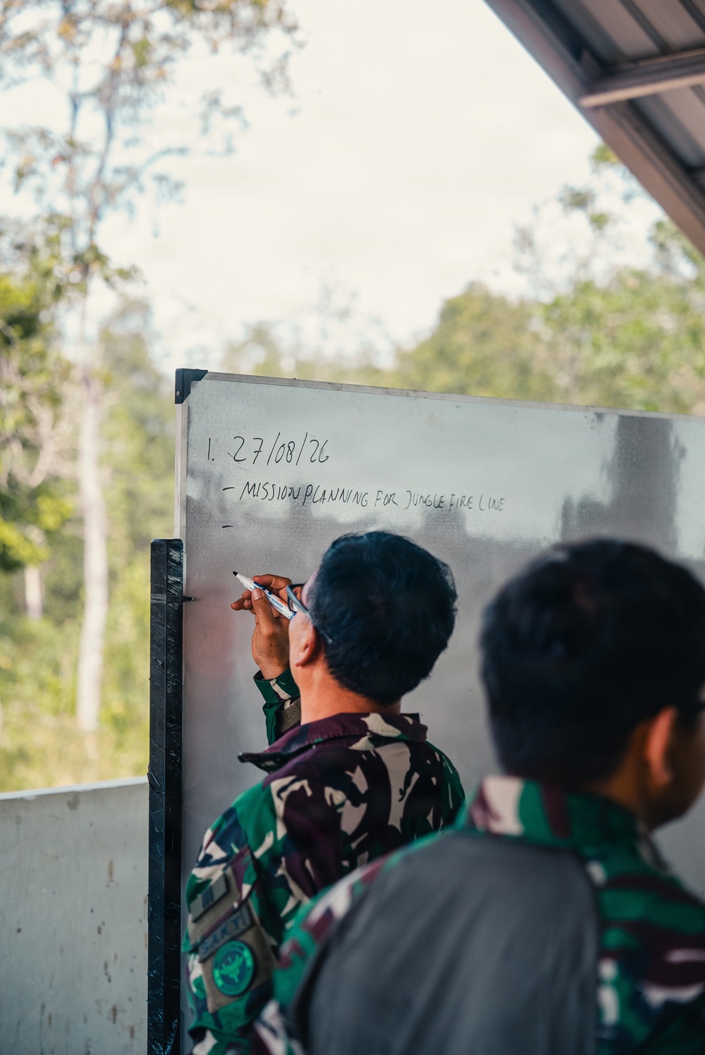 A person in military camouflage uniform is writing on a whiteboard with a marker. The whiteboard displays a date '27/08/26' and the text 'MISSION PLANNING FOR JUNGLE FIRE LINE'. A second individual in similar attire observes from the right foreground.