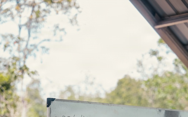 A person in military camouflage uniform is writing on a whiteboard with a marker. The whiteboard displays a date '27/08/26' and the text 'MISSION PLANNING FOR JUNGLE FIRE LINE'. A second individual in similar attire observes from the right foreground.