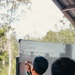 A person in military camouflage uniform is writing on a whiteboard with a marker. The whiteboard displays a date '27/08/26' and the text 'MISSION PLANNING FOR JUNGLE FIRE LINE'. A second individual in similar attire observes from the right foreground.