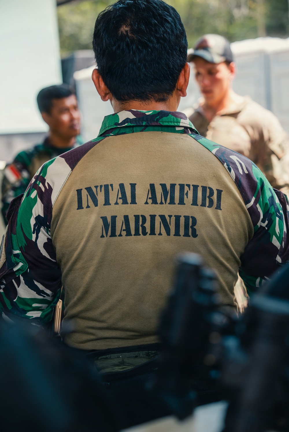 The image shows military personnel in a group setting, with the foreground figure wearing a distinctive Indonesian Marine Corps uniform. The back of the vest reads 'INTAI AMFIBI MARINIR', identifying the wearer as a member of the Indonesian Marine Amphibious Reconnaissance unit. At least one Western military personnel in tan/MultiCam uniform is visible in the background, suggesting a joint or multinational military exercise.