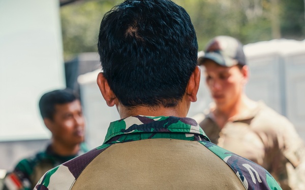 The image shows military personnel in a group setting, with the foreground figure wearing a distinctive Indonesian Marine Corps uniform. The back of the vest reads 'INTAI AMFIBI MARINIR', identifying the wearer as a member of the Indonesian Marine Amphibious Reconnaissance unit. At least one Western military personnel in tan/MultiCam uniform is visible in the background, suggesting a joint or multinational military exercise.
