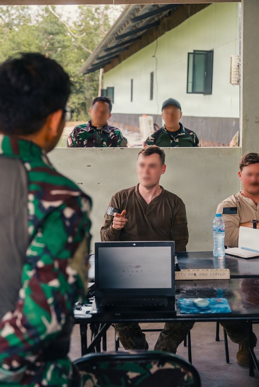 A military briefing or meeting is taking place in an open-air structure, with personnel from what appears to be multiple nations present. Two individuals in civilian/tactical clothing are seated at a table with a laptop, water bottle, and documents, while two soldiers in camouflage uniforms stand outside observing. A person in Indonesian TNI (Tentara Nasional Indonesia) camouflage uniform is visible in the foreground.