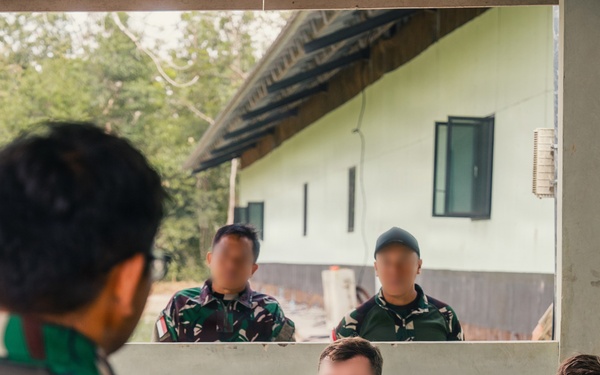 A military briefing or meeting is taking place in an open-air structure, with personnel from what appears to be multiple nations present. Two individuals in civilian/tactical clothing are seated at a table with a laptop, water bottle, and documents, while two soldiers in camouflage uniforms stand outside observing. A person in Indonesian TNI (Tentara Nasional Indonesia) camouflage uniform is visible in the foreground.