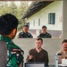 A military briefing or meeting is taking place in an open-air structure, with personnel from what appears to be multiple nations present. Two individuals in civilian/tactical clothing are seated at a table with a laptop, water bottle, and documents, while two soldiers in camouflage uniforms stand outside observing. A person in Indonesian TNI (Tentara Nasional Indonesia) camouflage uniform is visible in the foreground.