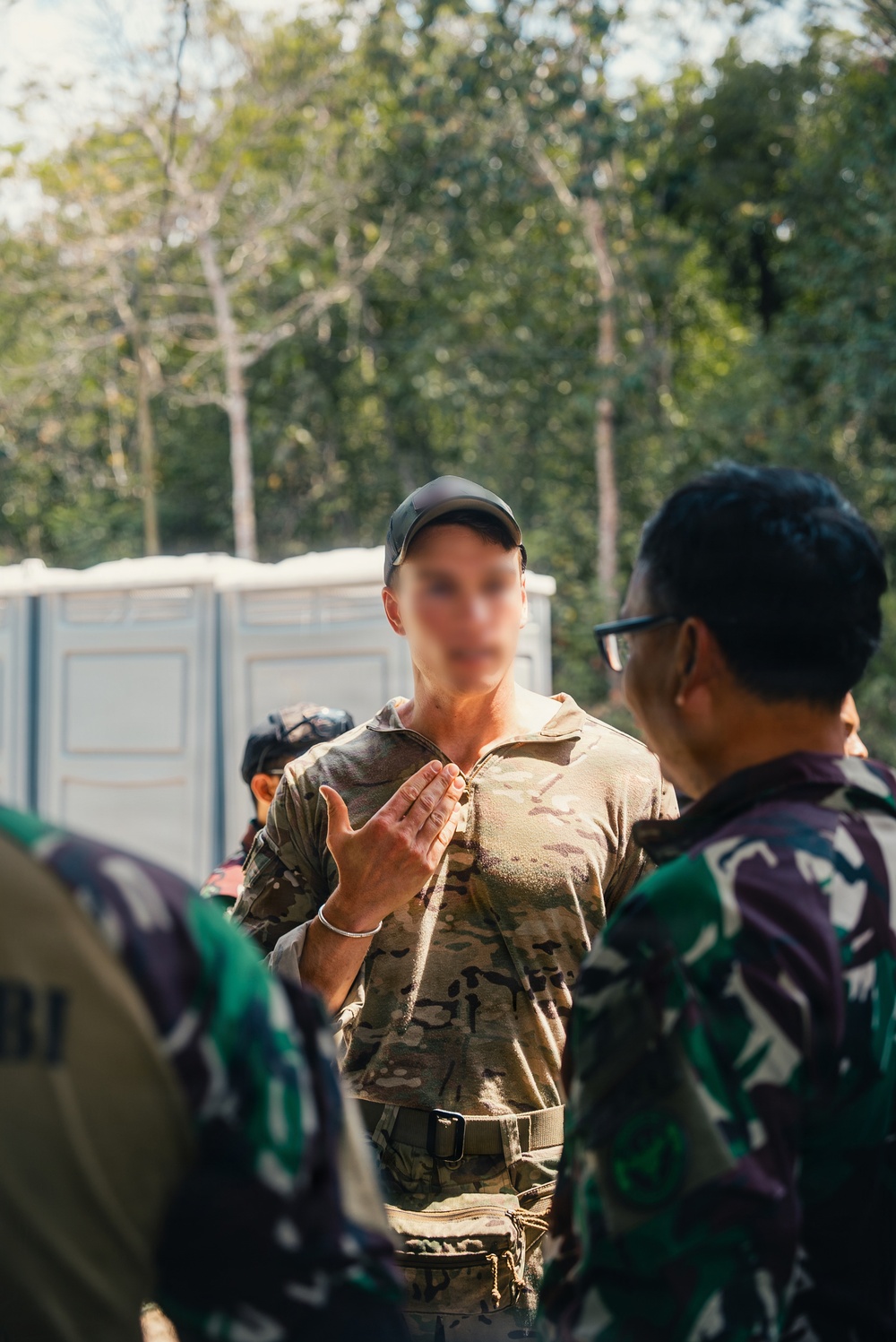 A military briefing or discussion is taking place outdoors among several personnel. A central figure wearing MultiCam/OCP pattern uniform and a dark baseball cap appears to be speaking and gesturing to a group. Other personnel wearing a different camouflage pattern (green/purple tones, consistent with Indonesian TNI uniform) are listening.