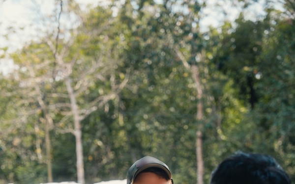 A military briefing or discussion is taking place outdoors among several personnel. A central figure wearing MultiCam/OCP pattern uniform and a dark baseball cap appears to be speaking and gesturing to a group. Other personnel wearing a different camouflage pattern (green/purple tones, consistent with Indonesian TNI uniform) are listening.