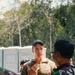 A military briefing or discussion is taking place outdoors among several personnel. A central figure wearing MultiCam/OCP pattern uniform and a dark baseball cap appears to be speaking and gesturing to a group. Other personnel wearing a different camouflage pattern (green/purple tones, consistent with Indonesian TNI uniform) are listening.