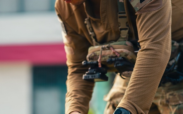 A military or tactical operator is handling what appears to be a parachute pack or specialized equipment bag. The individual is wearing multicam/OCP camouflage uniform with a tactical chest rig and a black digital watch. The image focuses on the hands and gear, with a blurred urban background.