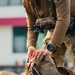 A military or tactical operator is handling what appears to be a parachute pack or specialized equipment bag. The individual is wearing multicam/OCP camouflage uniform with a tactical chest rig and a black digital watch. The image focuses on the hands and gear, with a blurred urban background.