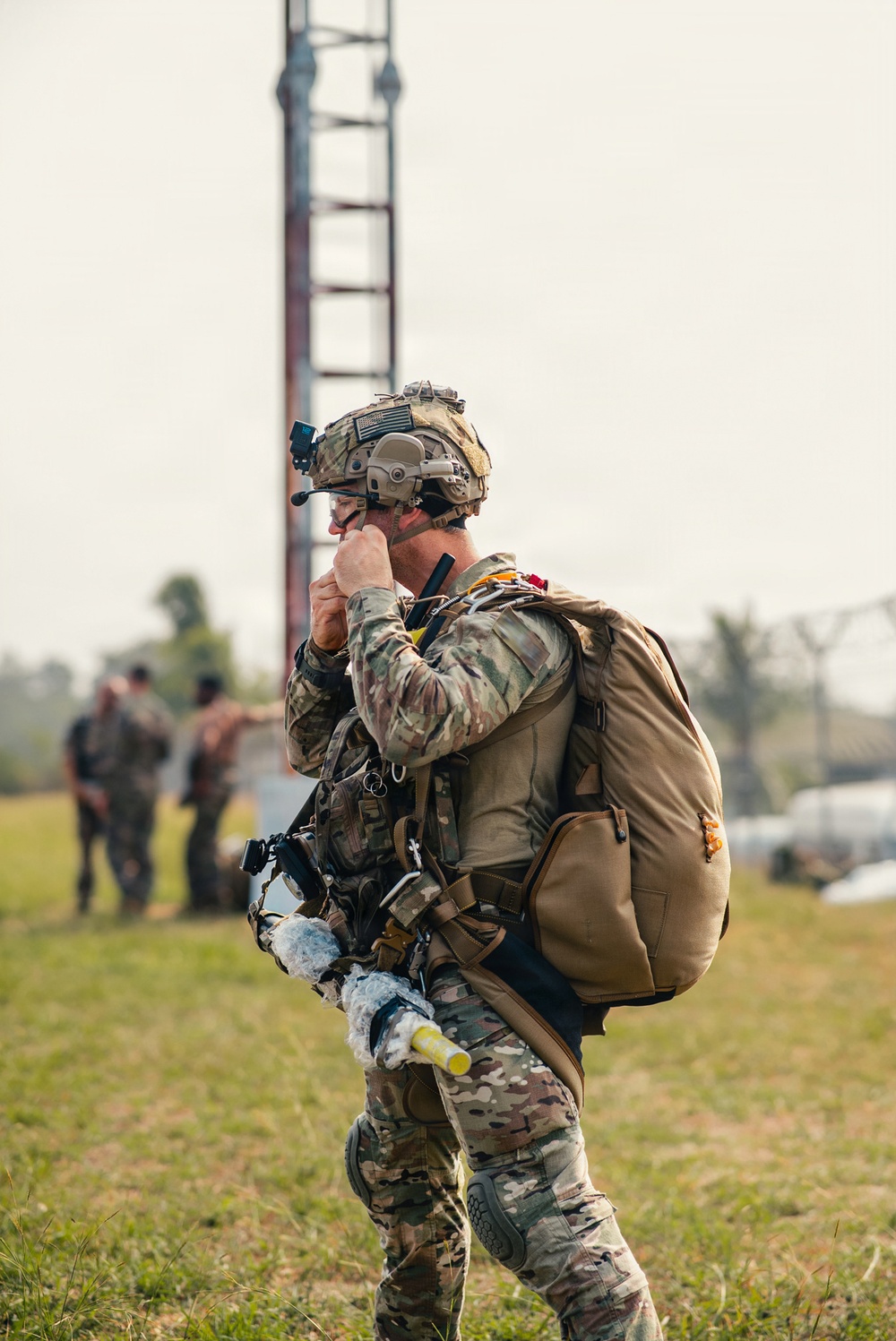 A heavily equipped soldier in MultiCam/OCP camouflage uniform is standing on a grassy field, holding a radio handset to his face while carrying a large rucksack. He appears to be communicating after what may be a parachute operation, evidenced by harness equipment and a partially visible parachute bag. Additional personnel are visible in the blurred background near a tall metal tower structure.