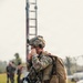 A heavily equipped soldier in MultiCam/OCP camouflage uniform is standing on a grassy field, holding a radio handset to his face while carrying a large rucksack. He appears to be communicating after what may be a parachute operation, evidenced by harness equipment and a partially visible parachute bag. Additional personnel are visible in the blurred background near a tall metal tower structure.
