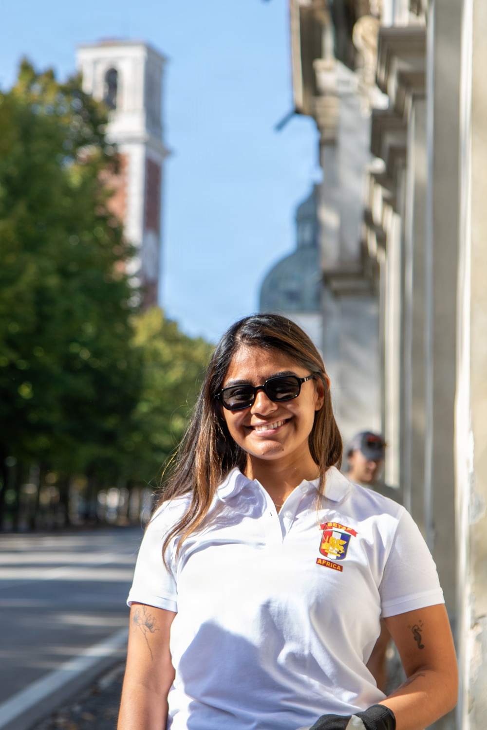 A smiling young woman wearing a white polo shirt with an embroidered badge and dark sunglasses stands on a sunny urban street. She has long dark hair with highlights and visible tattoos on both arms. The background features a tree-lined boulevard, a brick bell tower, and a domed building under a clear blue sky.
