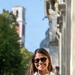 A smiling young woman wearing a white polo shirt with an embroidered badge and dark sunglasses stands on a sunny urban street. She has long dark hair with highlights and visible tattoos on both arms. The background features a tree-lined boulevard, a brick bell tower, and a domed building under a clear blue sky.