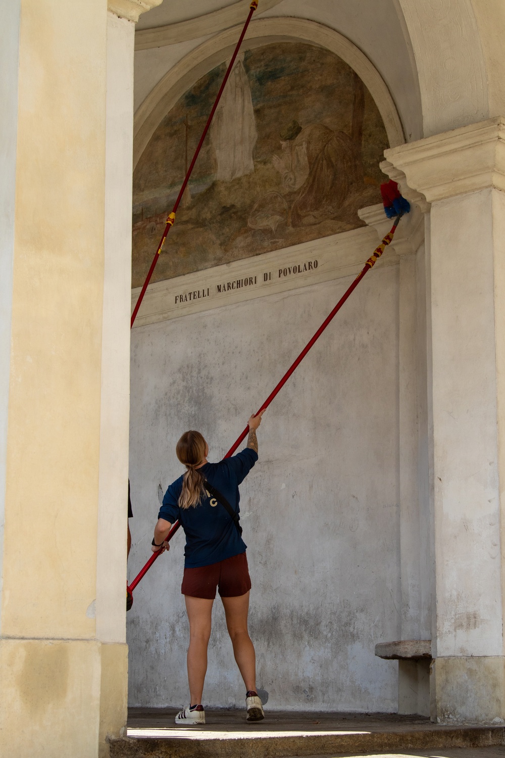 A young woman with blonde hair in a ponytail is using a long red extension pole with a duster attachment to clean or dust the upper architectural elements of what appears to be an Italian religious or historic building. The arched niche behind her contains a deteriorated fresco depicting religious figures, with an inscription reading 'FRATELLI MARCHIORI DI POVOLARO' on the molding below it. The setting appears to be an outdoor portico or loggia with classical architectural details.