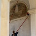 A young woman with blonde hair in a ponytail is using a long red extension pole with a duster attachment to clean or dust the upper architectural elements of what appears to be an Italian religious or historic building. The arched niche behind her contains a deteriorated fresco depicting religious figures, with an inscription reading 'FRATELLI MARCHIORI DI POVOLARO' on the molding below it. The setting appears to be an outdoor portico or loggia with classical architectural details.
