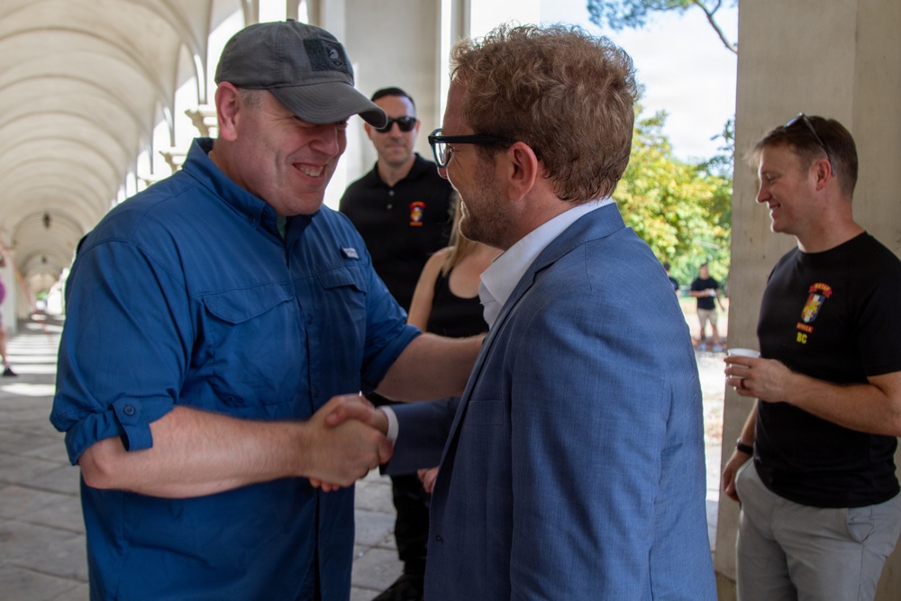 Two men are shaking hands in what appears to be a colonnaded outdoor walkway or portico. One man wears a blue utility shirt and gray baseball cap, while the other wears a light blue blazer and black-framed glasses. Several other individuals are visible in the background, including two men wearing black shirts with a logo reading 'DC'.