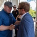 Two men are shaking hands in what appears to be a colonnaded outdoor walkway or portico. One man wears a blue utility shirt and gray baseball cap, while the other wears a light blue blazer and black-framed glasses. Several other individuals are visible in the background, including two men wearing black shirts with a logo reading 'DC'.