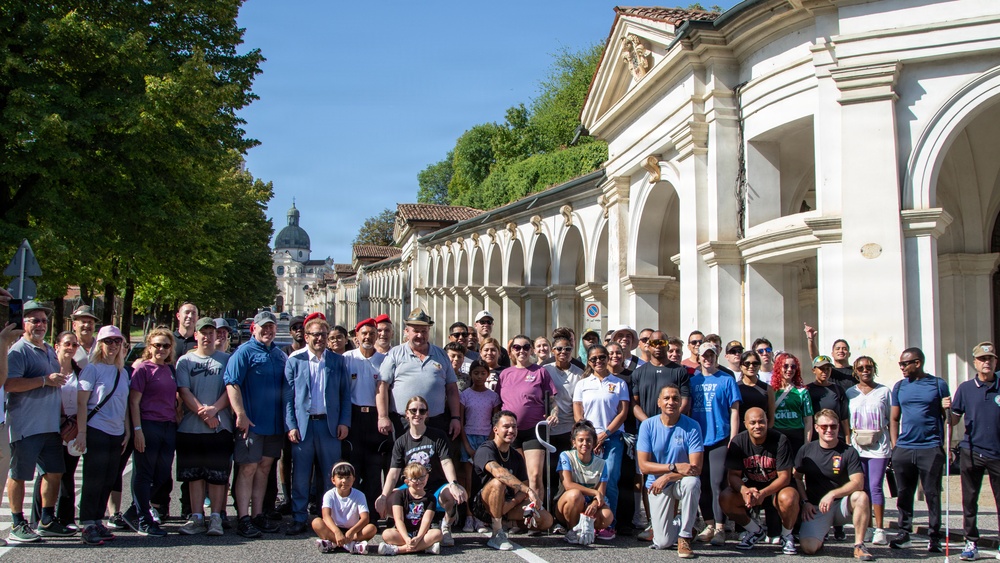 A large group of approximately 50-60 people poses for a group photograph on a street in what appears to be an Italian city, likely Vicenza, based on the classical Palladian-style arcaded architecture visible in the background. The group appears diverse in age and ethnicity, dressed in casual clothing, suggesting a community or organizational event such as a walk or tour. A domed church or basilica is visible in the background along a tree-lined avenue.