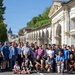 A large group of approximately 50-60 people poses for a group photograph on a street in what appears to be an Italian city, likely Vicenza, based on the classical Palladian-style arcaded architecture visible in the background. The group appears diverse in age and ethnicity, dressed in casual clothing, suggesting a community or organizational event such as a walk or tour. A domed church or basilica is visible in the background along a tree-lined avenue.