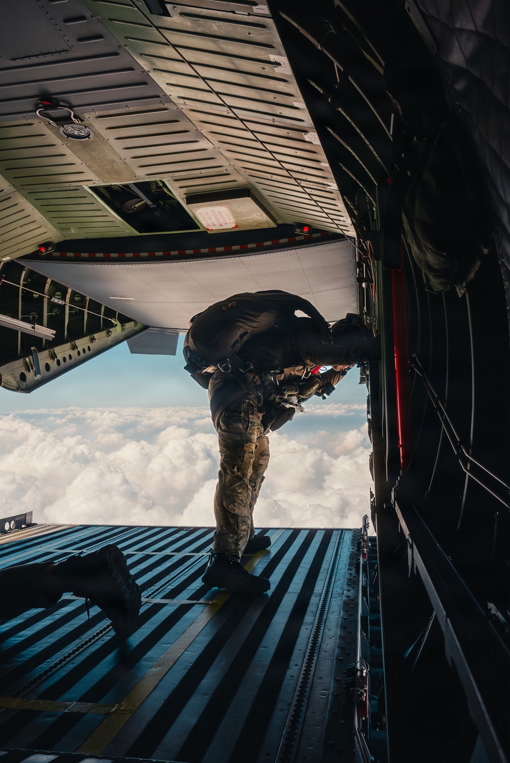 A military paratrooper in full combat gear is positioned at the open rear ramp of a large military cargo aircraft, appearing to be at the moment of or preparing for a jump. The aircraft is at high altitude above a cloud layer, with bright daylight visible through the opening. The image is taken from inside the aircraft looking toward the ramp exit.