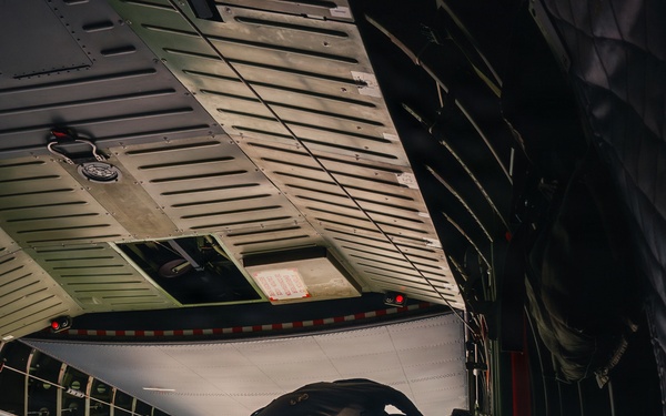 A military paratrooper in full combat gear is positioned at the open rear ramp of a large military cargo aircraft, appearing to be at the moment of or preparing for a jump. The aircraft is at high altitude above a cloud layer, with bright daylight visible through the opening. The image is taken from inside the aircraft looking toward the ramp exit.