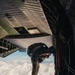 A military paratrooper in full combat gear is positioned at the open rear ramp of a large military cargo aircraft, appearing to be at the moment of or preparing for a jump. The aircraft is at high altitude above a cloud layer, with bright daylight visible through the opening. The image is taken from inside the aircraft looking toward the ramp exit.