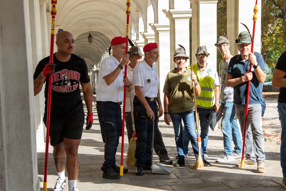 A group of men are gathered under a colonnaded arcade, holding long-handled brooms and cleaning tools, apparently participating in a community cleanup event. Several men are wearing distinctive red berets and traditional Alpine-style hats with feathers, suggesting possible affiliation with a veterans or civic organization. The setting appears to be a historic building or public monument in what looks like an Italian city.
