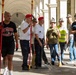 A group of men are gathered under a colonnaded arcade, holding long-handled brooms and cleaning tools, apparently participating in a community cleanup event. Several men are wearing distinctive red berets and traditional Alpine-style hats with feathers, suggesting possible affiliation with a veterans or civic organization. The setting appears to be a historic building or public monument in what looks like an Italian city.