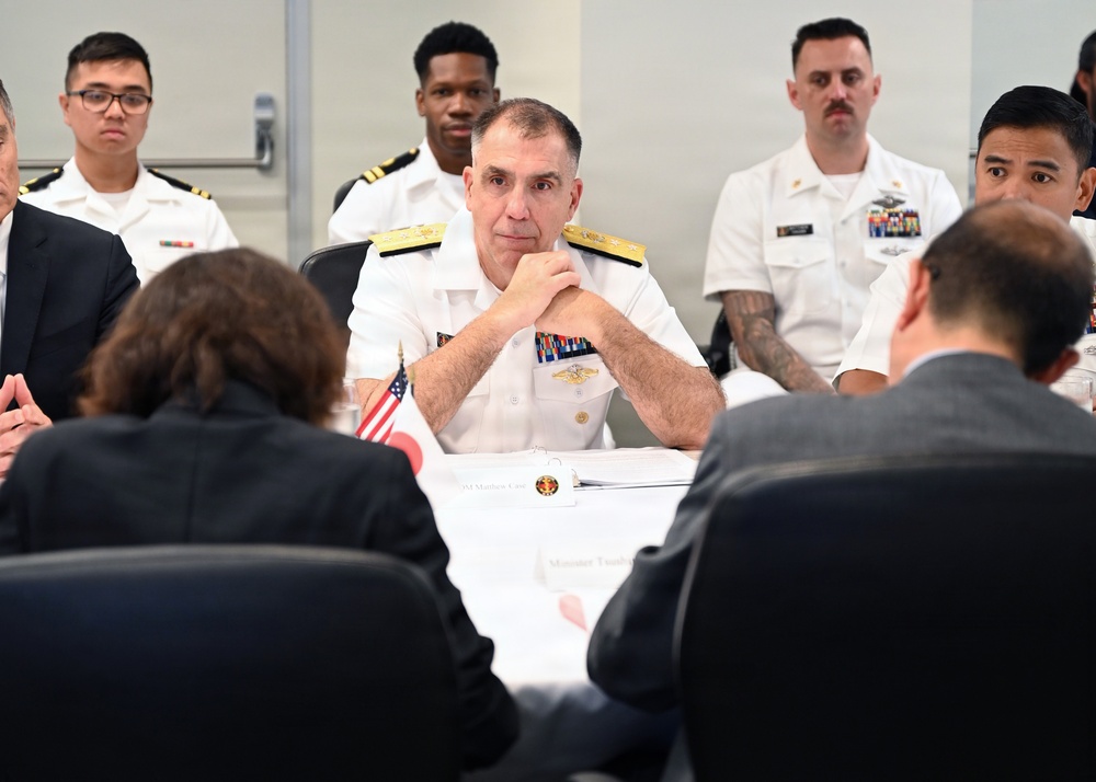 A formal meeting or briefing is taking place at a conference table, with a senior U.S. Navy admiral as the central figure, flanked by military personnel in white dress uniforms and civilians in dark suits. Small American and Japanese flags are visible on the table, suggesting a bilateral or diplomatic military meeting. The admiral appears attentive, with hands clasped, listening to individuals seated across from him.