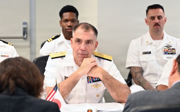 A formal meeting or briefing is taking place at a conference table, with a senior U.S. Navy admiral as the central figure, flanked by military personnel in white dress uniforms and civilians in dark suits. Small American and Japanese flags are visible on the table, suggesting a bilateral or diplomatic military meeting. The admiral appears attentive, with hands clasped, listening to individuals seated across from him.