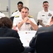 A formal meeting or briefing is taking place at a conference table, with a senior U.S. Navy admiral as the central figure, flanked by military personnel in white dress uniforms and civilians in dark suits. Small American and Japanese flags are visible on the table, suggesting a bilateral or diplomatic military meeting. The admiral appears attentive, with hands clasped, listening to individuals seated across from him.